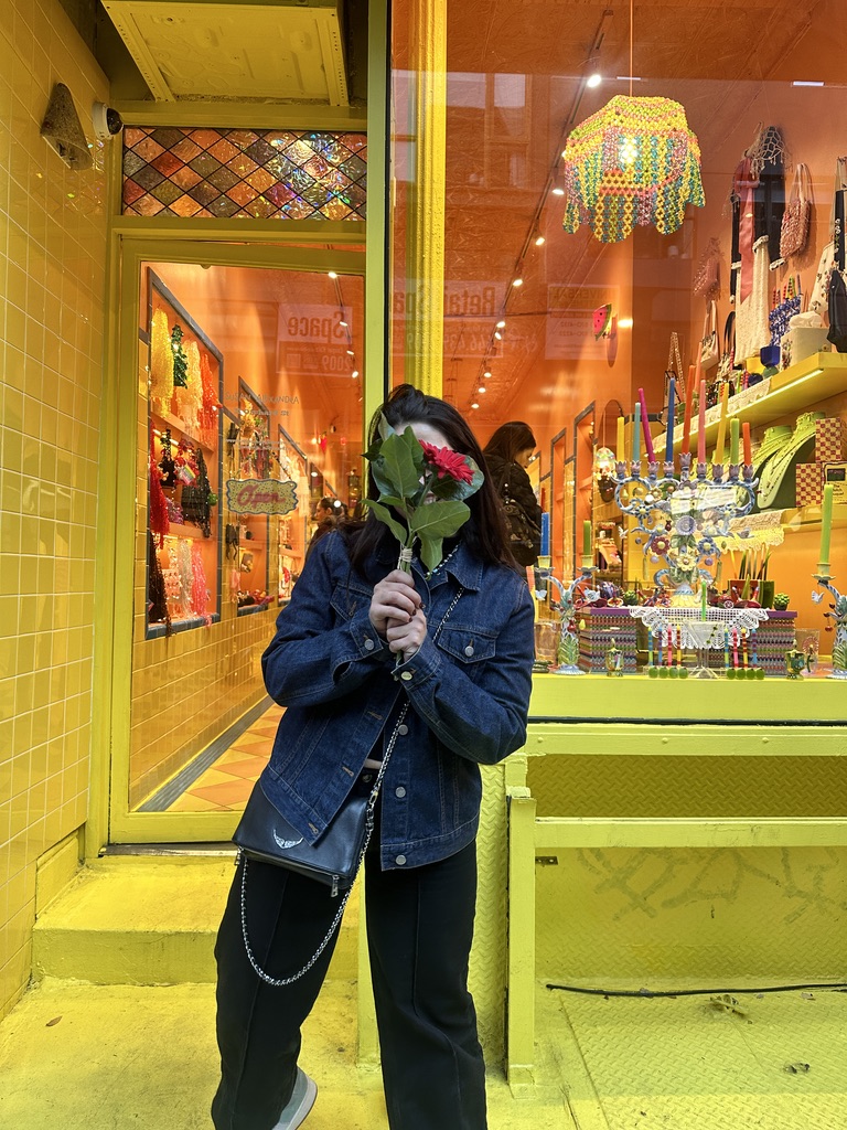 Annette frente a una tienda amarilla, tapándose la cara con una flor roja