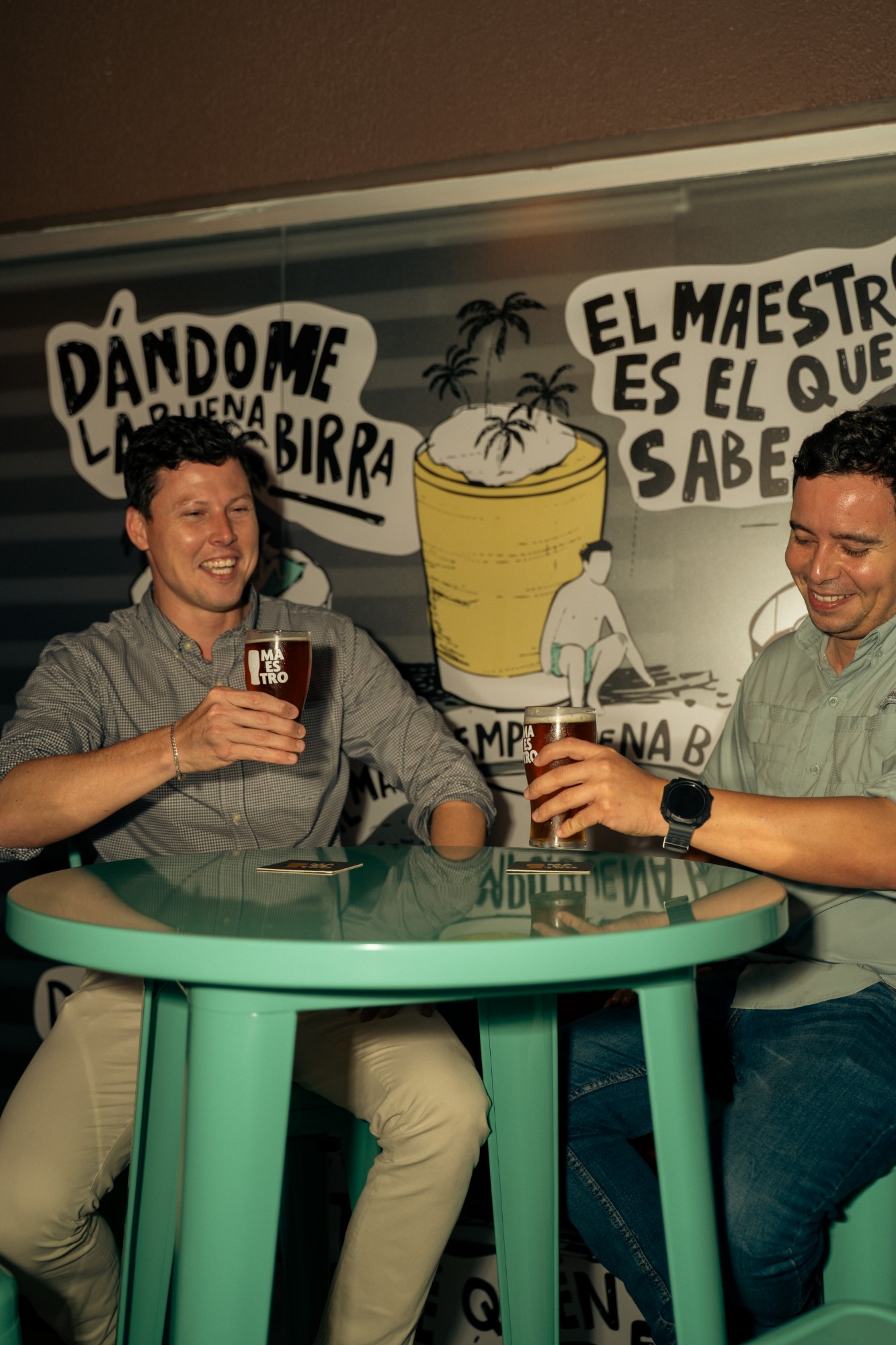 Dos personas brindando frente a un mural ilustrado de Maestro